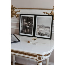 Load image into Gallery viewer, Two framed fine art prints, one with Paris rooftops, other with cloud in palace with chandeliers, next to each other on antique table with gold details against ornamental wall with gold details, champagne glass and cork also on table, Paris, French interior
