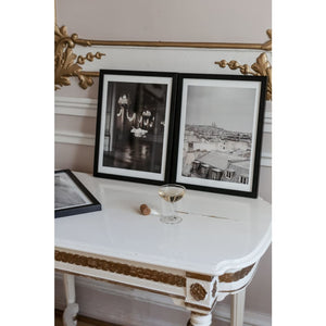 Two framed fine art prints, one with Paris rooftops, other with cloud in palace with chandeliers, next to each other on antique table with gold details against ornamental wall with gold details, champagne glass and cork also on table, Paris, French interior