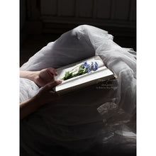 Load image into Gallery viewer, close up of bottom half of person lying on an old wooden veranda floor, hands holding an old book with freshly picked blue forget-me-not flowers, tulle skirt with light sifting through the hem, softness, fine art print
