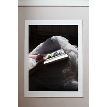 Load image into Gallery viewer, close up of bottom half of person lying on an old wooden veranda floor, hands holding an old book with freshly picked blue forget-me-not flowers, tulle skirt with light sifting through the hem, softness, fine art print with white border on pale wall

